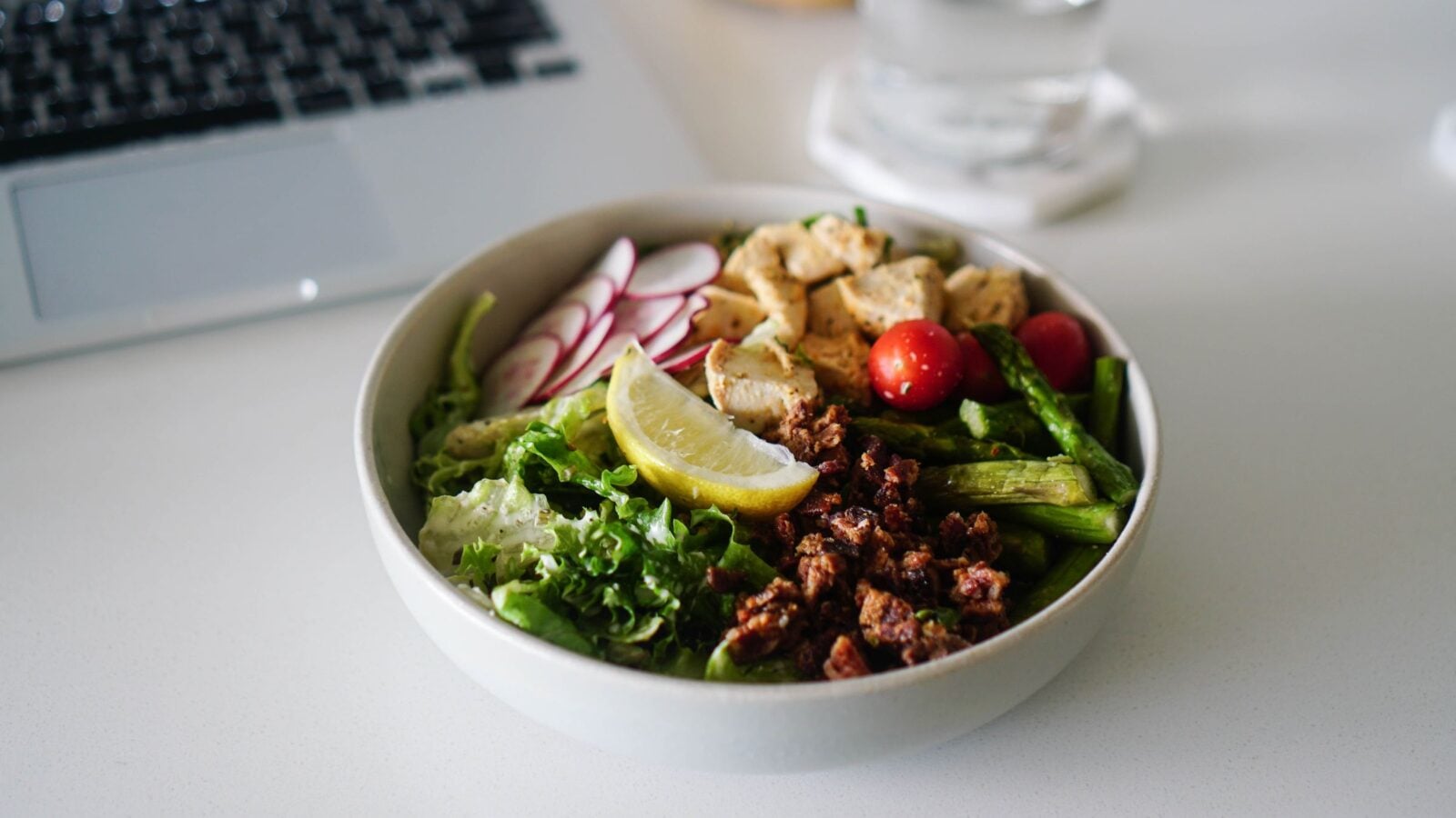 A colorful lunch bowl of vegetables and protein beside a laptop