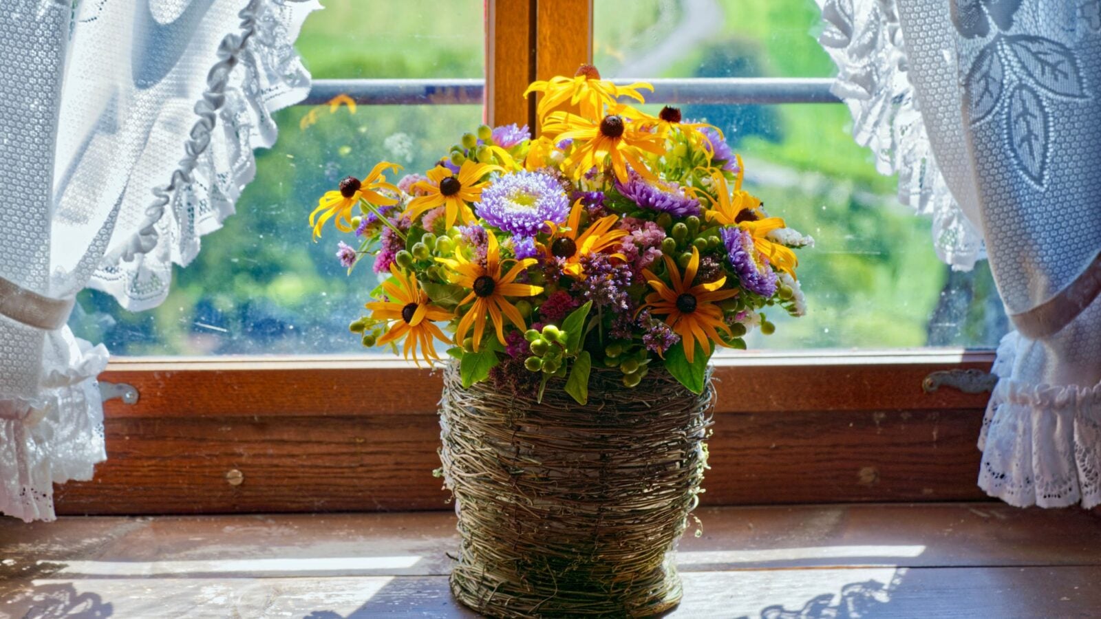 A basket of wildflowers on a windowsill with lace curtains